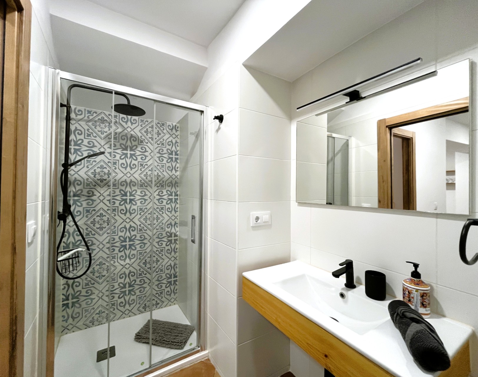 Modernes Badezimmer mit begehbarer Dusche, dekorativen Fliesen und schwarzen Armaturen. Waschtisch mit Holzakzenten und ein großer Spiegel mit linearer Beleuchtung.