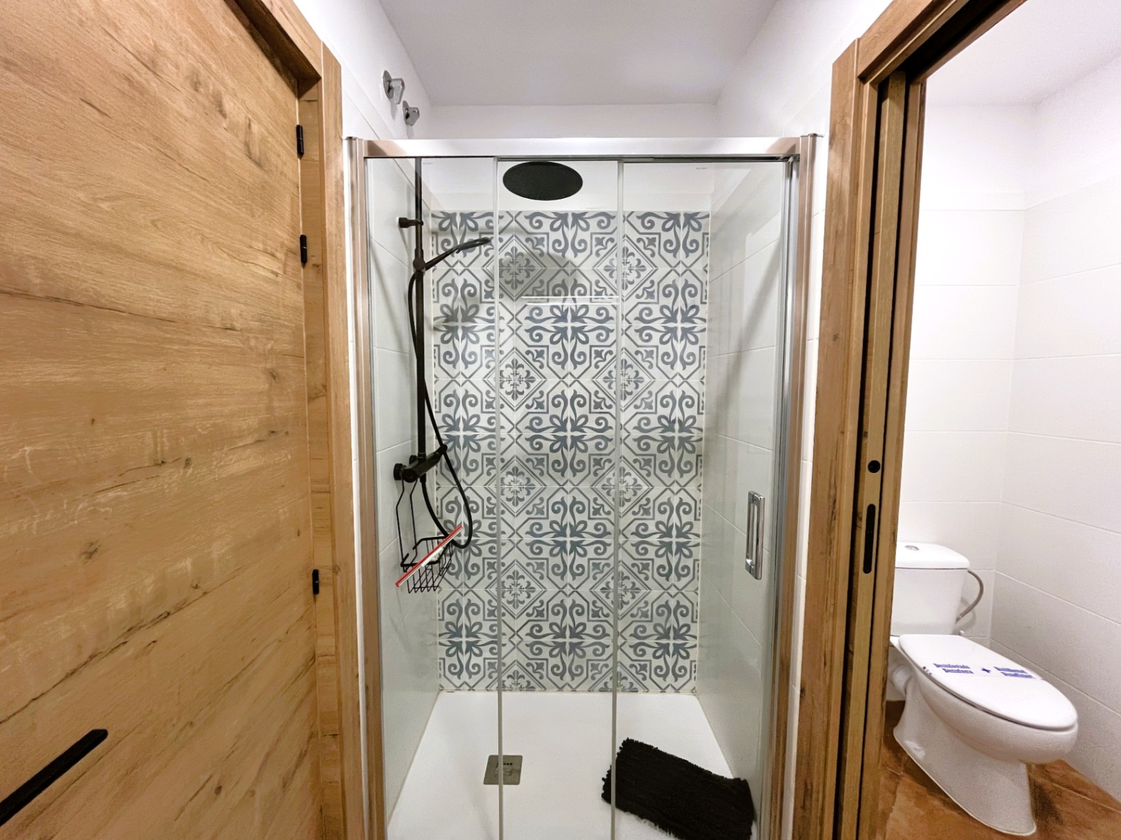 Modernisiertes Badezimmer mit begehbarer Dusche, Glastüren und dekorativen Fliesen im Retro-Stil. Schwarze Armaturen und Türen in natürlicher Holzoptik.