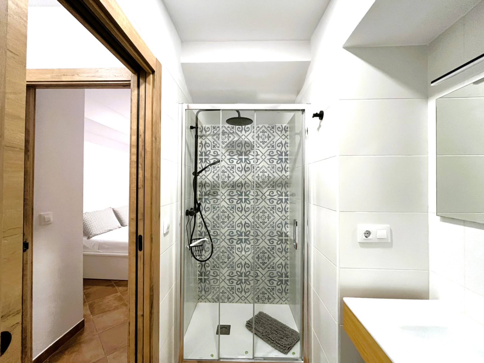 Modernisiertes Badezimmer mit ebenerdiger Dusche, dekorativen Musterfliesen und schwarzen Armaturen. Blick durch den Holzrahmen in das helle Schlafzimmer.