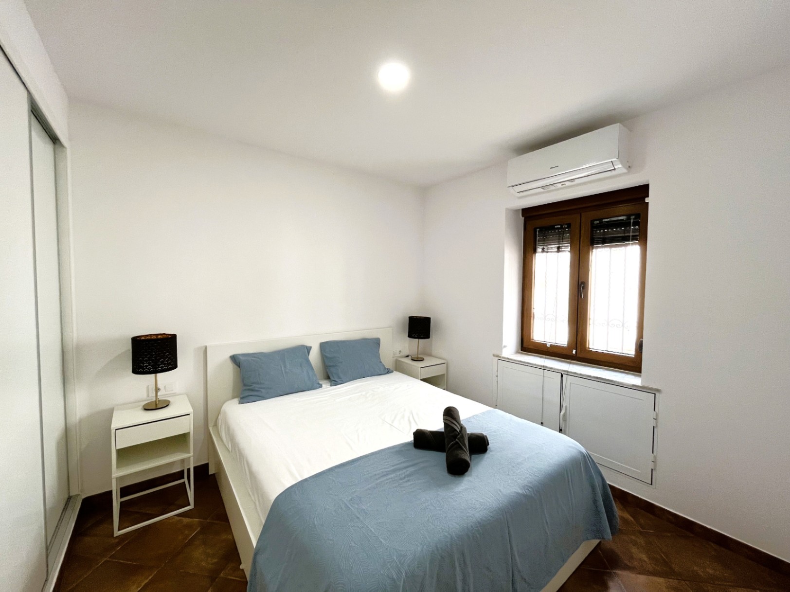 Schlafzimmer mit Doppelbett, weißen Nachttischen und Klimaanlage. Ausgestattet mit rustikalem Fliesenboden, Einbauschrank und Holzfenster mit Jalousien.