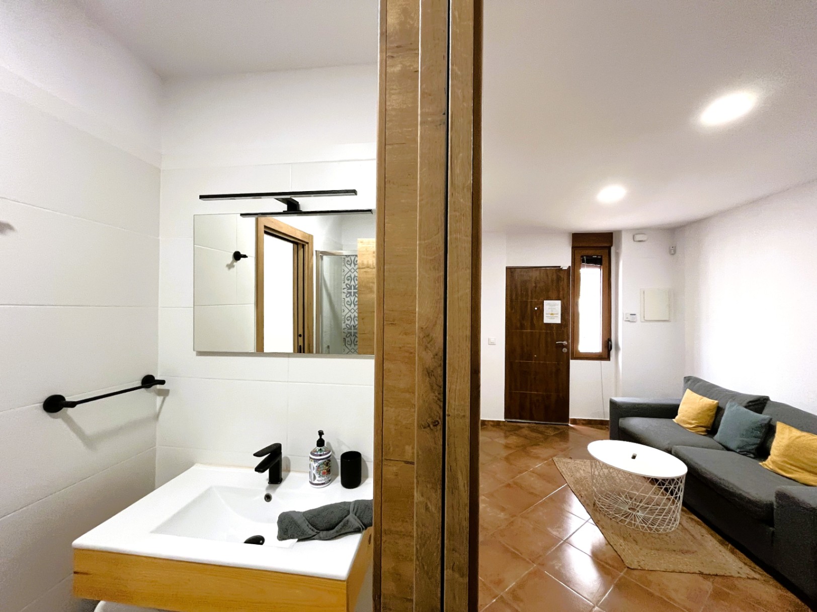 Wohnbereich mit Terrakottaboden und grauem Sofa neben einem modernen Badezimmer mit weißem Waschbecken, schwarzen Armaturen und hellen Wandfliesen.
