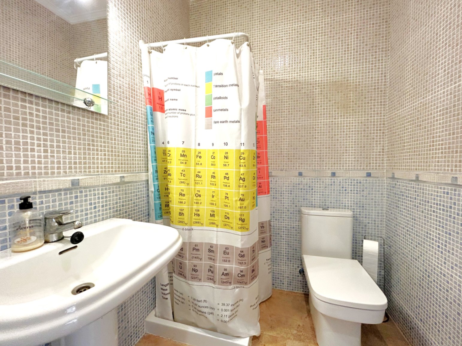 Badezimmer mit zweifarbigen Mosaikfliesen, weißem Säulenwaschbecken und Duschvorhang mit Periodensystem. Ausgestattet mit einem modernen WC und einem Spiegel mit Glasablage.
