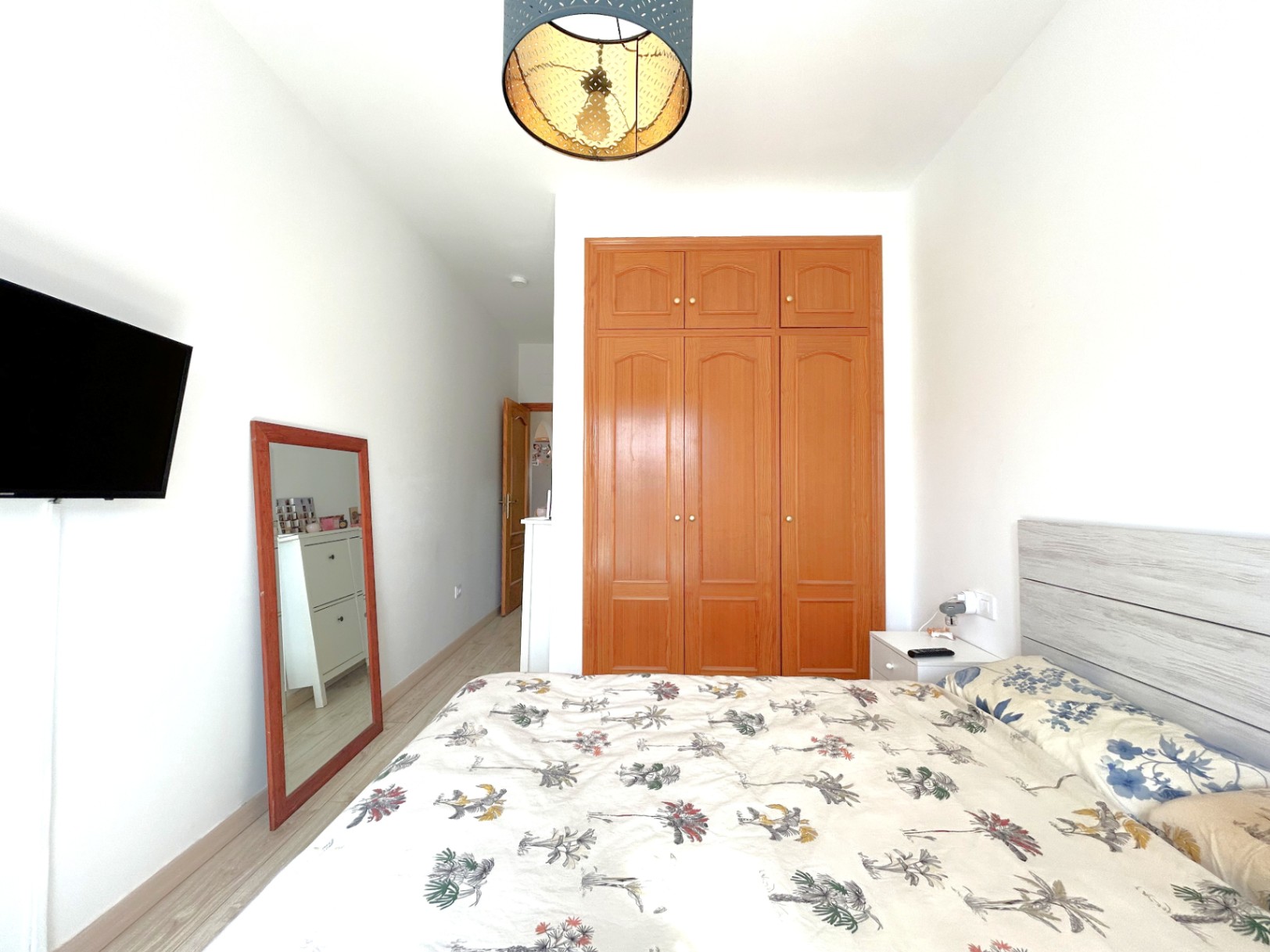 Doppelzimmer mit Einbauschrank aus Holz, hellem Laminatboden und wandmontiertem Fernseher. Ein Ganzkörperspiegel und eine dekorative Deckenleuchte ergänzen den Raum.