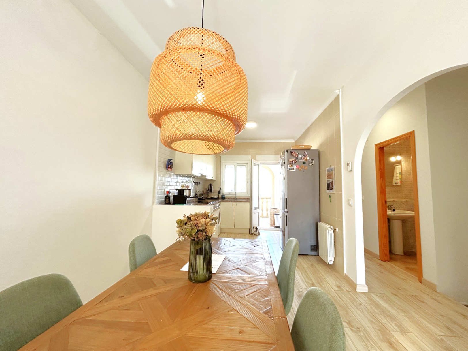 Essbereich mit markantem Holztisch und Korbgeflecht-Leuchte, integriert in eine offene Küche mit weißen Fronten und hellem Laminatboden.