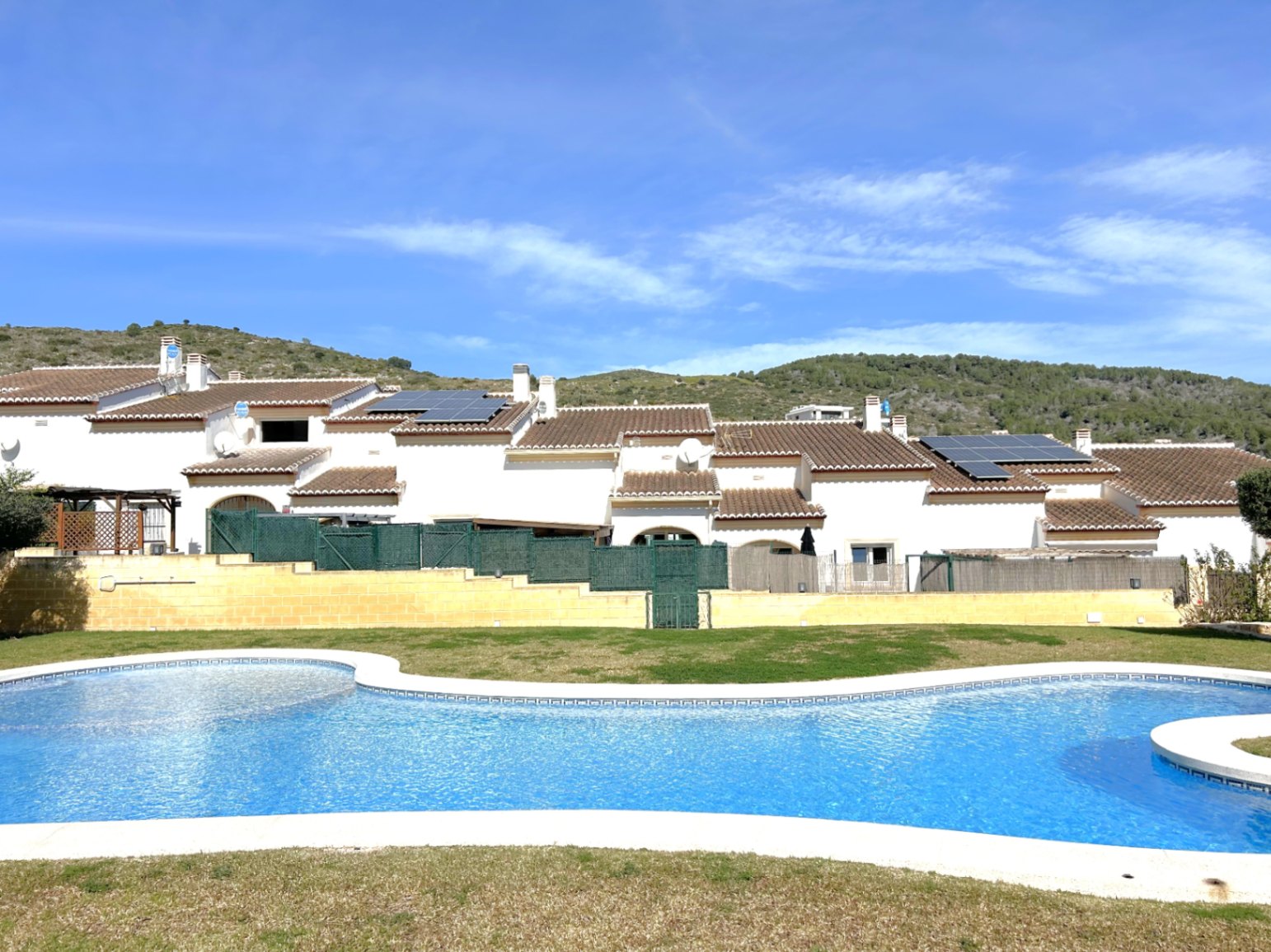 Großer Gemeinschaftspool mit geschwungenen Formen und Liegewiese, im Hintergrund die weißen Reihenhäuser mit Solarpaneelen und Blick auf die Hügel.