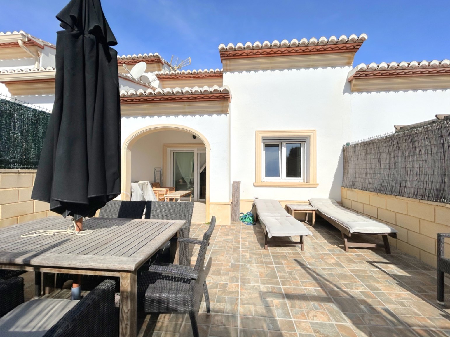Private, geflieste Terrasse mit Essbereich aus Holz, Sonnenliegen und einem schattigen Rundbogen-Eingang. Mediterrane Fassade mit klassischen Dachziegeln.