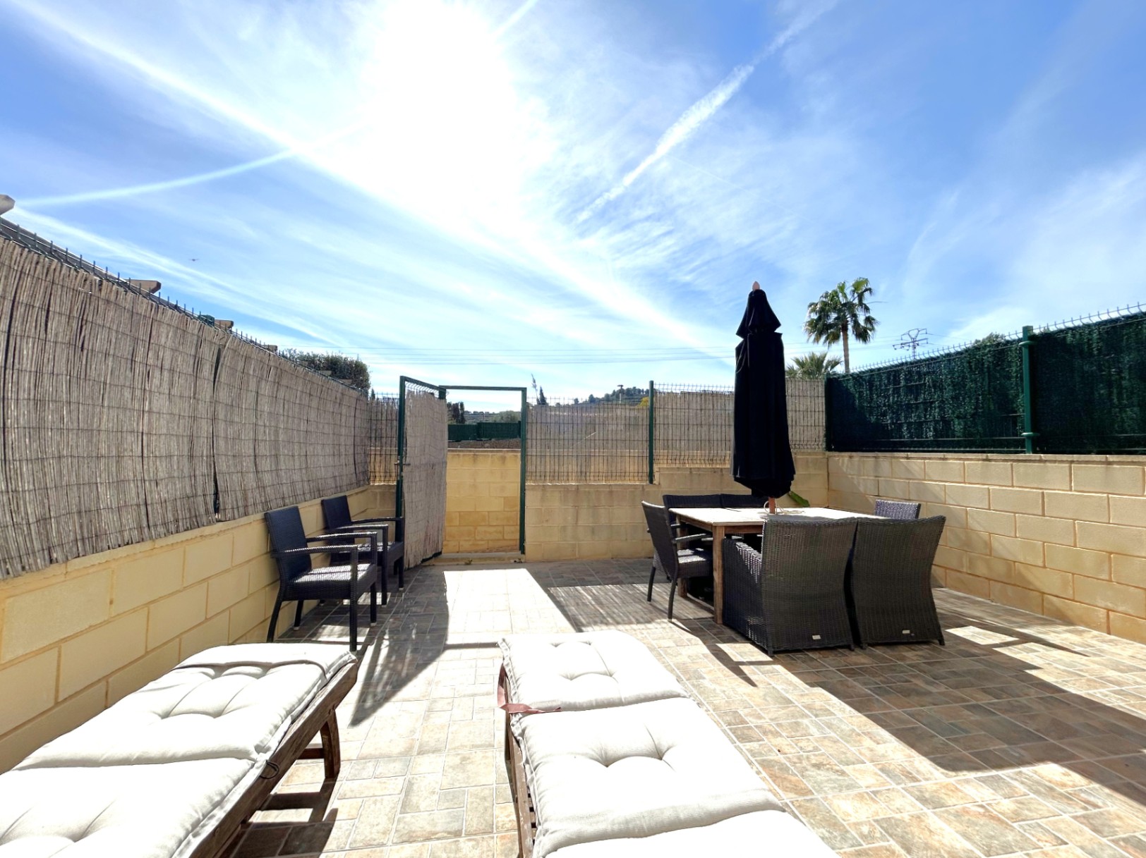 Südterrasse mit Steinzeugfliesen, ausgestattet mit einem Essbereich im Freien, Sonnenliegen und einem Sichtschutz aus Naturrohr für maximale Privatsphäre.