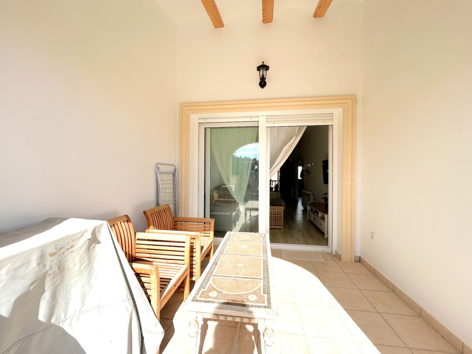 Überdachte Terrasse mit sichtbaren Holzbalken, Keramikfliesen und Holzmöbeln. Die Glasschiebetür ermöglicht einen direkten Übergang zum Wohnbereich.