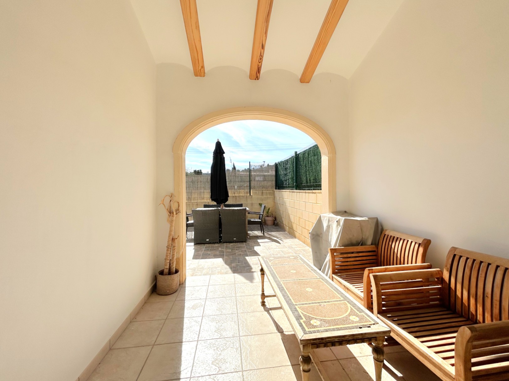 Überdachte Terrasse mit sichtbaren Holzbalken und Rundbogen, ausgestattet mit Holzmöbeln und direktem Zugang zum sonnigen Außenbereich.
