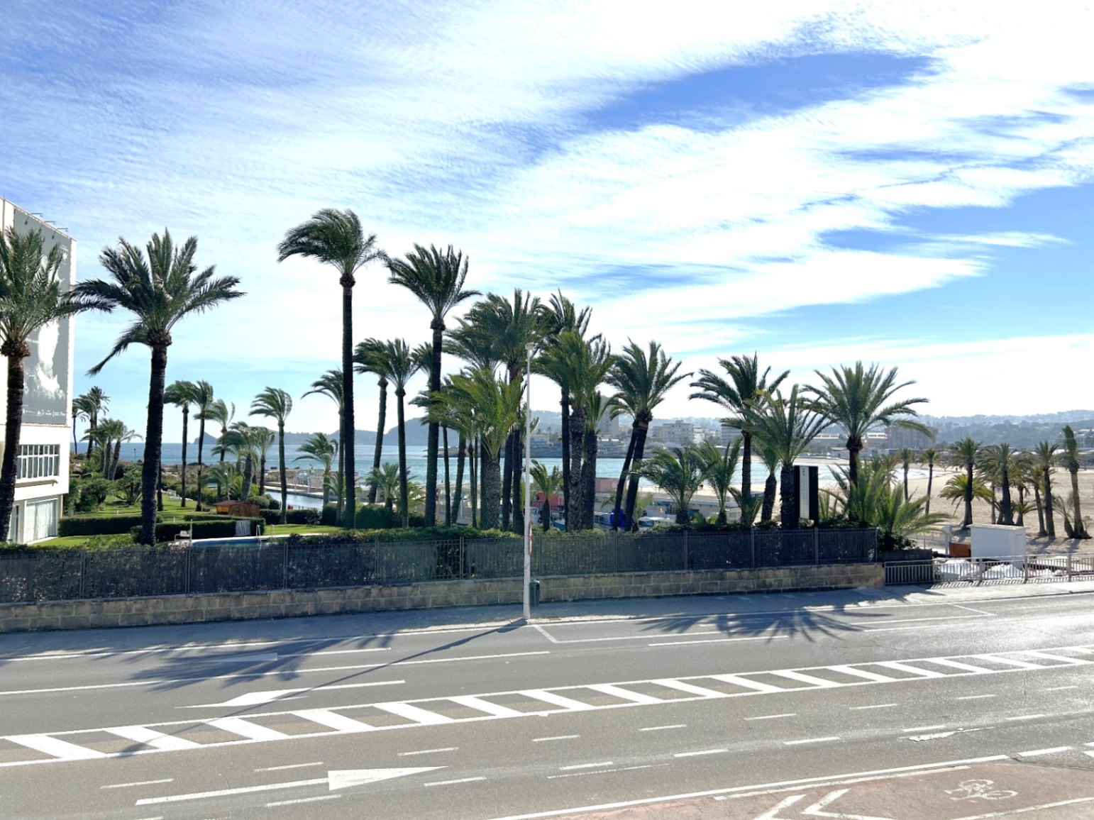 Erhöhter Blick von der Terrasse auf eine Küstenstraße, eine Reihe von Palmen und das Mittelmeer unter leicht bewölktem Himmel.