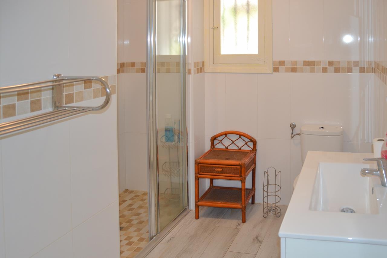 Badezimmer mit weiß gekachelten Wänden und Mosaikbordüre. Ausgestattet mit Glasduschkabine, integriertem Waschbecken und einem Beistelltisch aus Korbgeflecht.