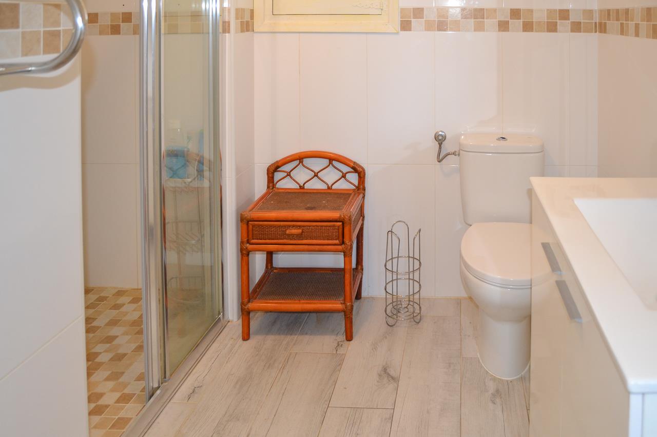 Funktionales Badezimmer mit Glasdusche, weißem WC und Rattantisch. Hell geflieste Wände mit Mosaikbordüre treffen auf einen Bodenbelag in heller Holzoptik.