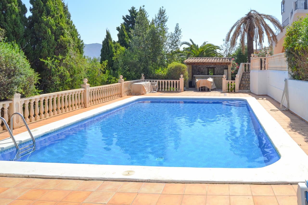 Großzügiger 12x6 Meter Pool mit blauen Mosaikfliesen, umgeben von einer weitläufigen Terrasse mit Steinbalustrade und einer überdachten Außenbar im privaten, grünen Garten.
