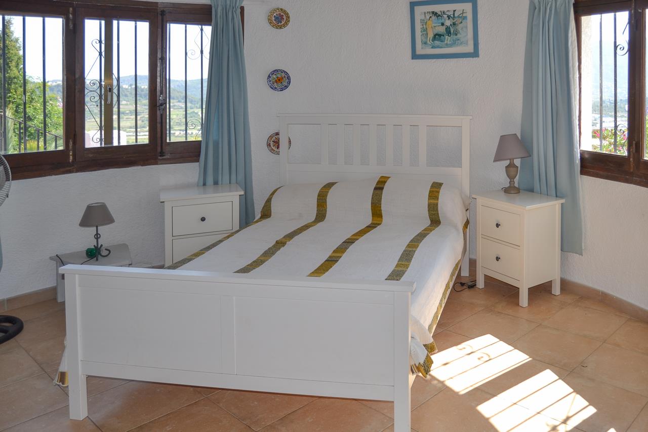 Helles Schlafzimmer mit weißem Holzbett, Terrakottaboden und Fenstern, die einen weiten Blick über das Tal von Pedreguer bieten.