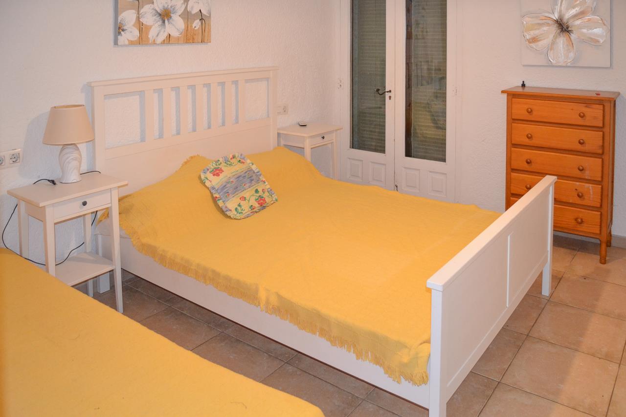 Helles Schlafzimmer mit weißem Holzbettrahmen, passenden Nachttischen und einer Kommode aus Naturholz. Fliesenboden und direkter Zugang nach draußen.