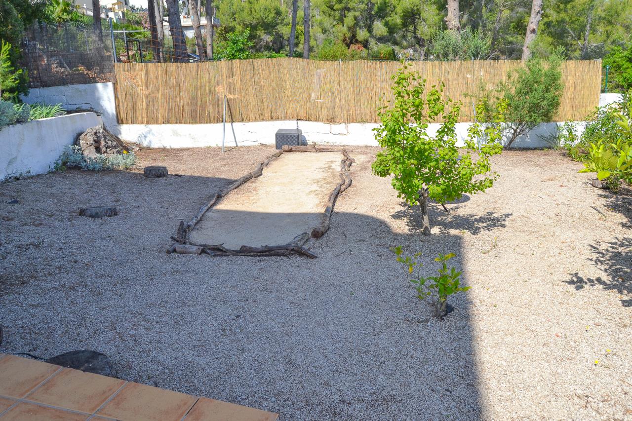 Pflegeleichter Kiesgarten mit einem rustikalen, von Naturstämmen eingefassten Bouleplatz, umgeben von Sichtschutz aus Schilfrohr und mediterranen Pflanzen.