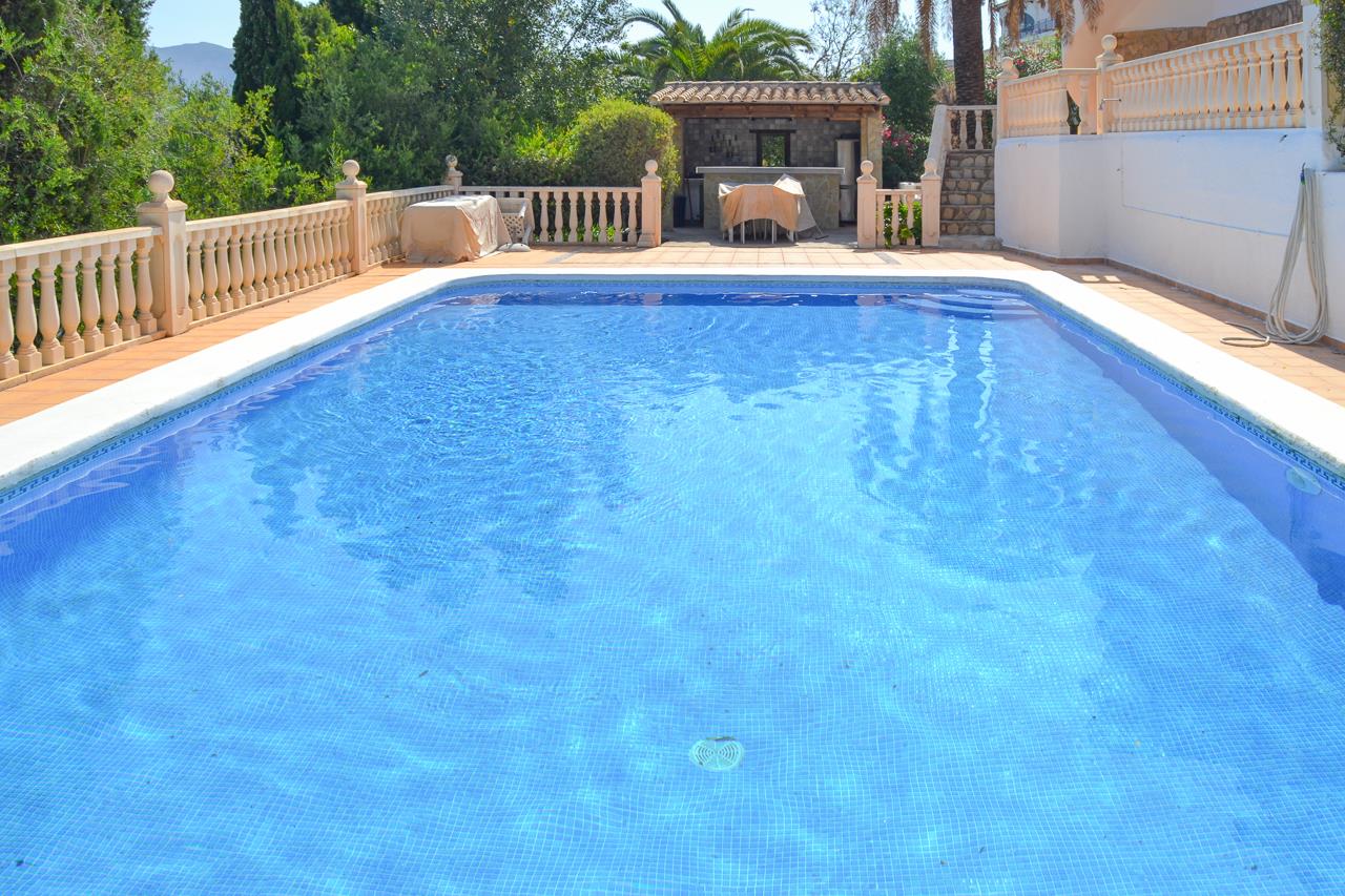 Rechteckiger Pool mit blauem Mosaik, umgeben von einer großzügigen Terrasse mit Balustraden und einem überdachten Außenbar-Bereich im Hintergrund.