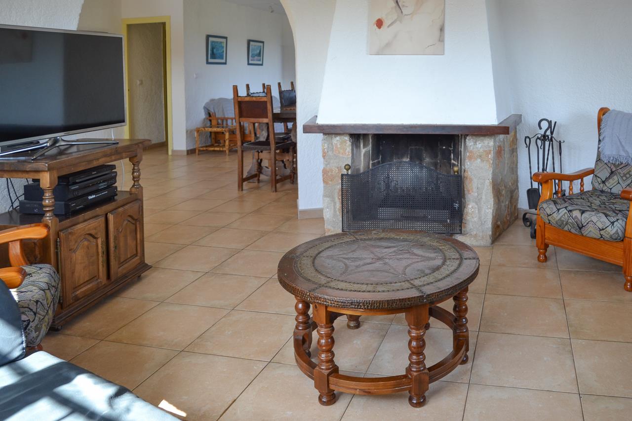 Rustikales Wohnzimmer mit Natursteinkamin, Fliesenboden und traditionellen Holzmöbeln. Ein Rundbogen verbindet den Raum mit dem angrenzenden Essbereich.