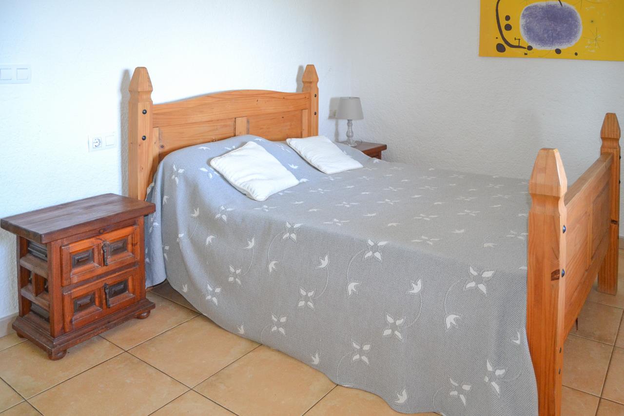 Schlafzimmer mit massivem Holzbettrahmen, rustikalem Nachttisch und Fliesenboden. Strukturierte weiße Wände unterstreichen die traditionelle Einrichtung und das natürliche Licht.