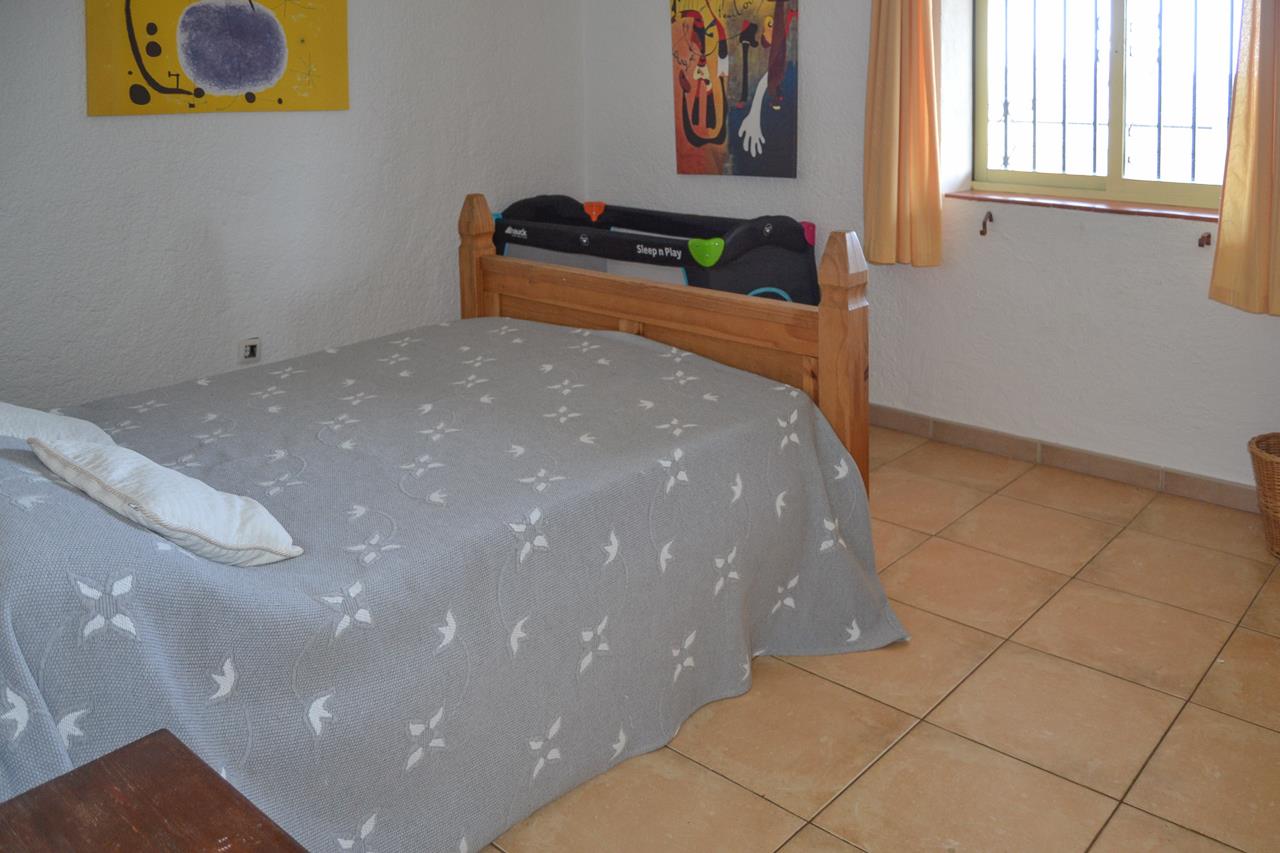 Schlafzimmer mit rustikalen Fliesenböden, Massivholz-Bettgestell und Reisekinderbett. Die weißen Wände sind strukturiert und das Fenster ist mit Gittern versehen.