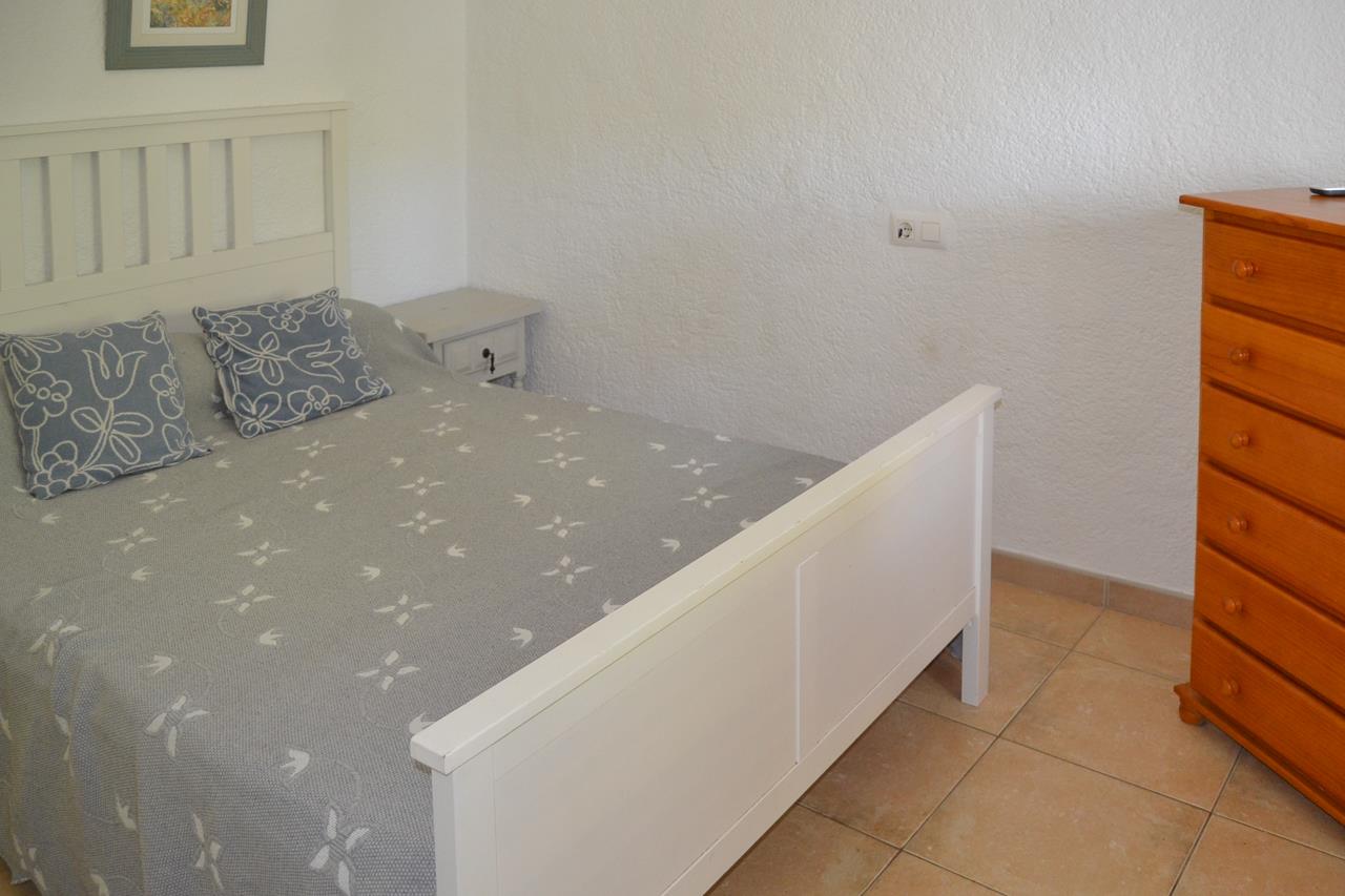 Schlafzimmer mit weißem Holzbett, Terrakotta-Fliesenboden und einer hohen Holzkommode vor hellen, strukturierten Wänden.