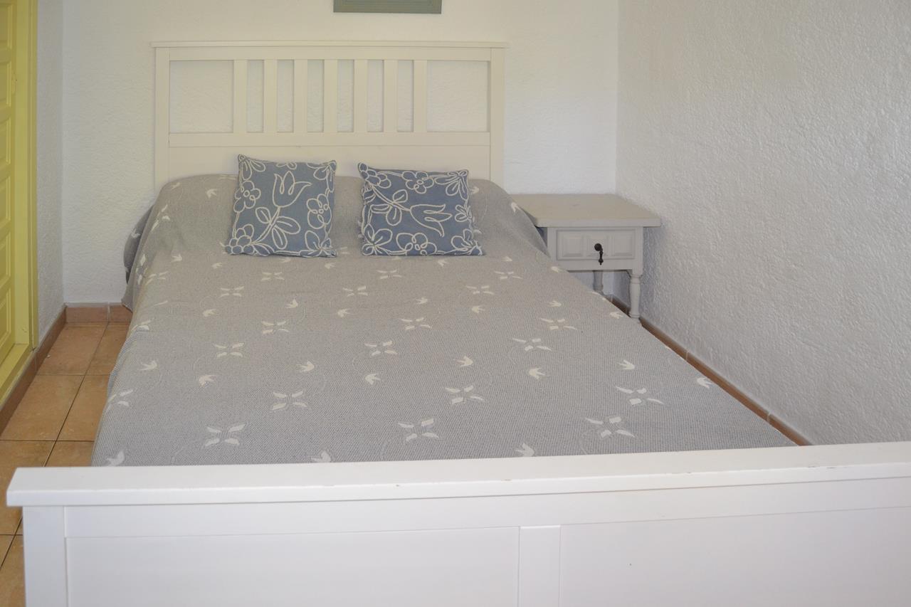 Schlafzimmer mit weißem Holzbettrahmen, passendem Nachttisch und Fliesenboden. Strukturierte weiße Wände und Bettwäsche in Grau- und Blautönen.
