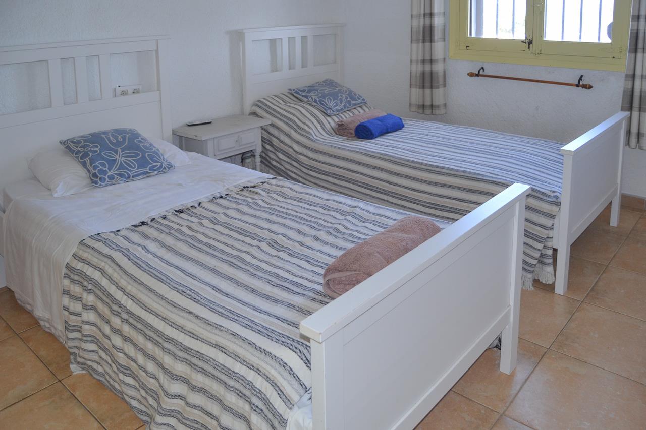Schlafzimmer mit zwei weißen Holzeinzelbetten, rustikalem Fliesenboden und Fenster mit gelbem Rahmen und karierten Vorhängen.