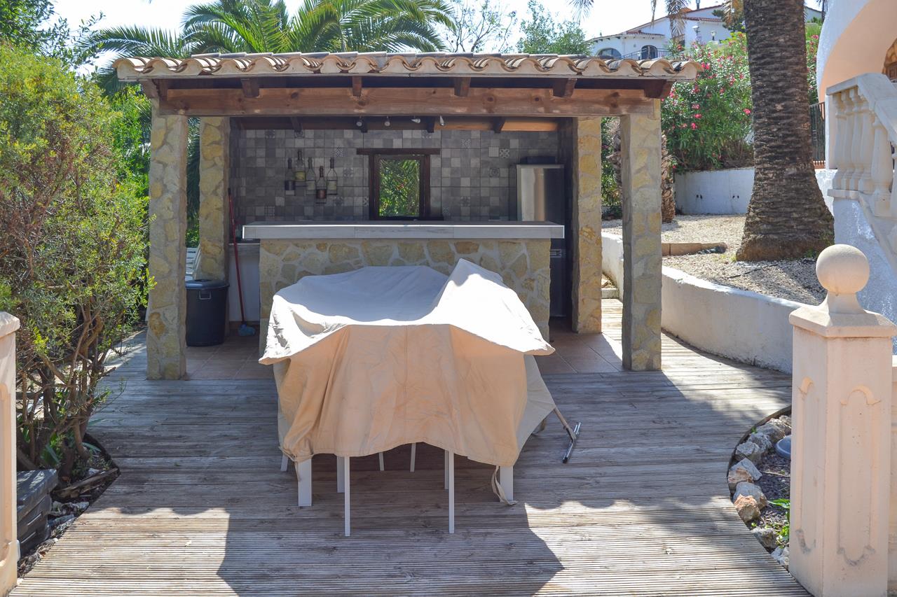 Überdachte Außenküche mit Steinbar, Holzbalkendecke und Holzterrasse, umgeben von mediterraner Bepflanzung und Palmen.