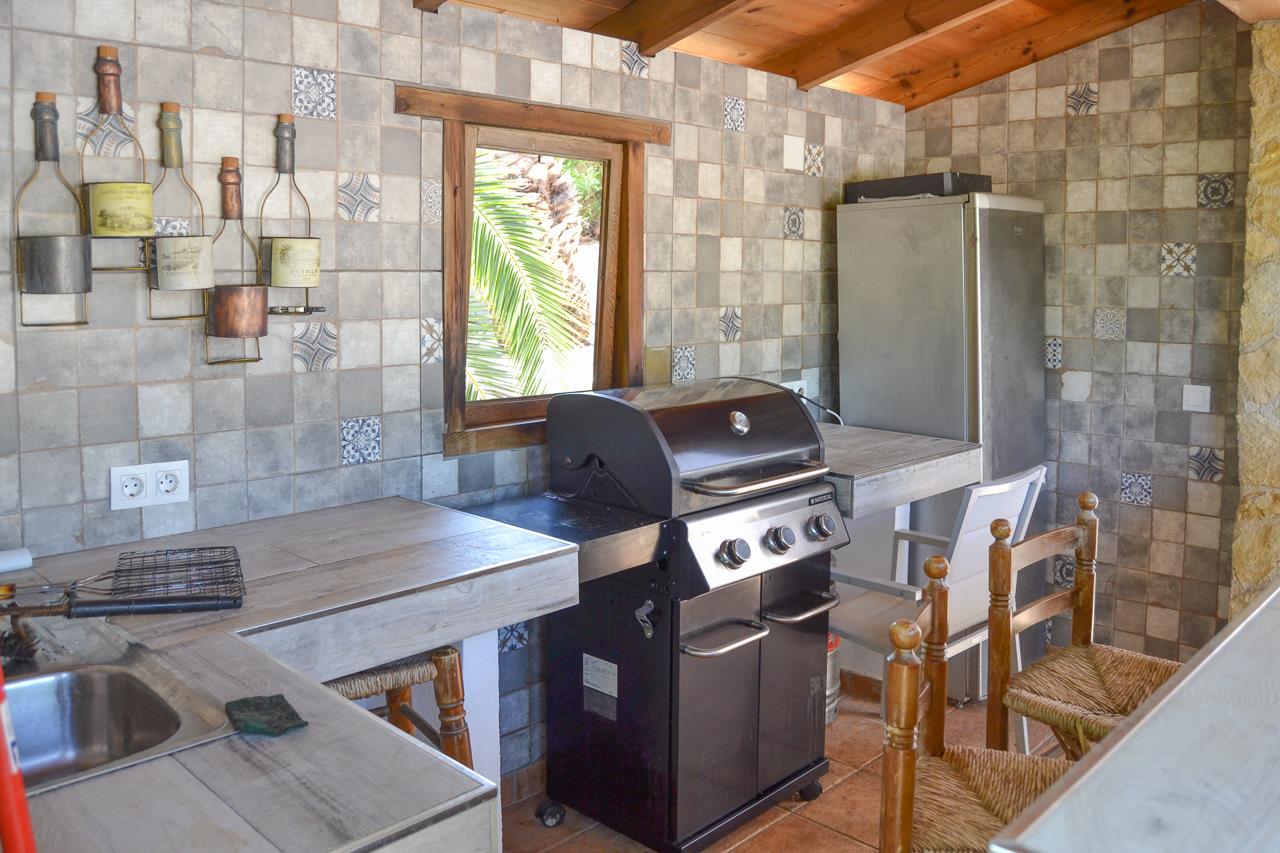 Überdachte Sommerküche mit Gasgrill, gefliesten Arbeitsplatten und rustikalen Wandmosaiken. Mit Holzbalkendecke, Spülbecken und Blick auf die umliegende Vegetation.