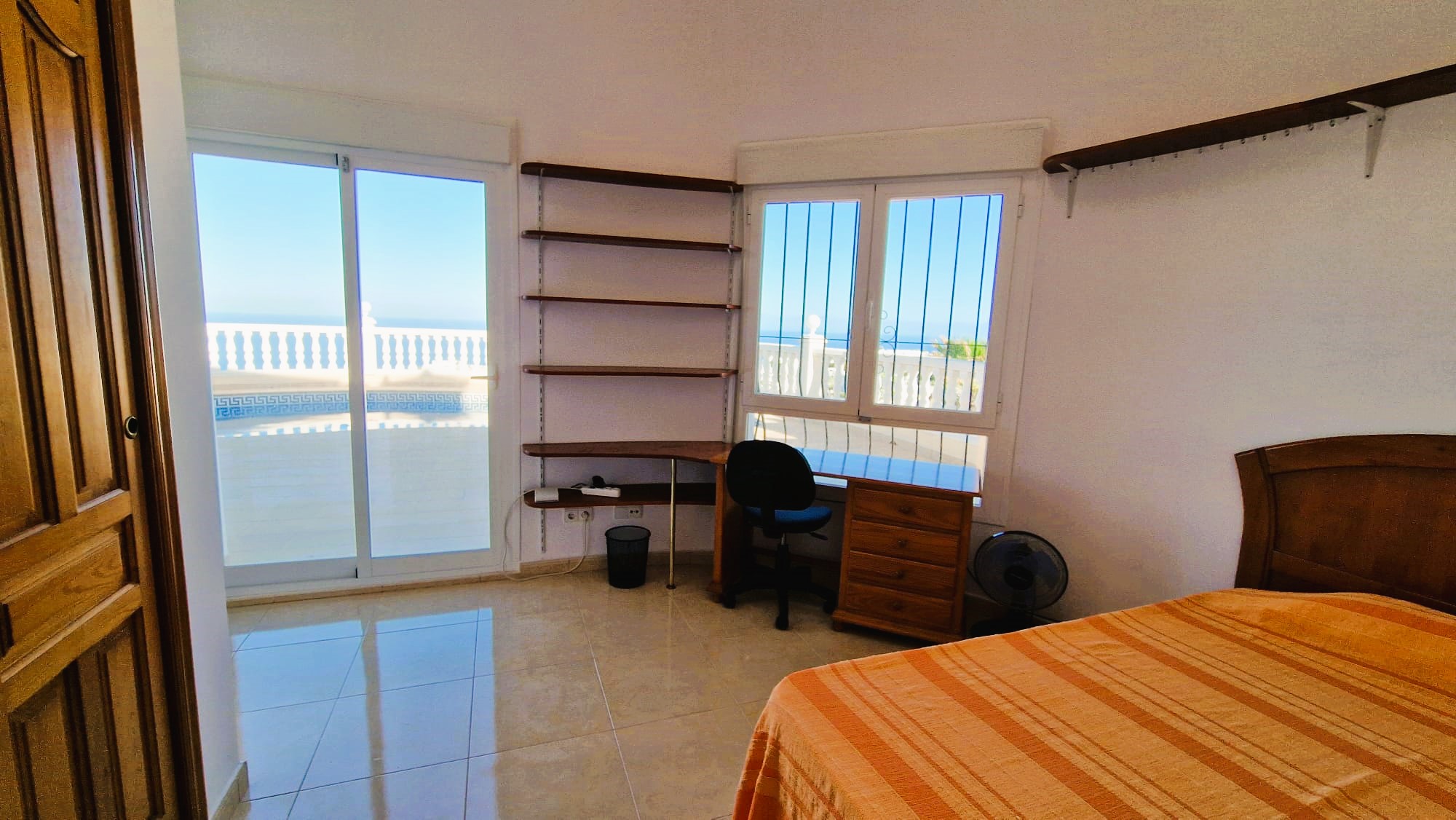 Helles Schlafzimmer mit Holzschreibtisch, Regalen und Blick auf den Balkon mit weißem Geländer.