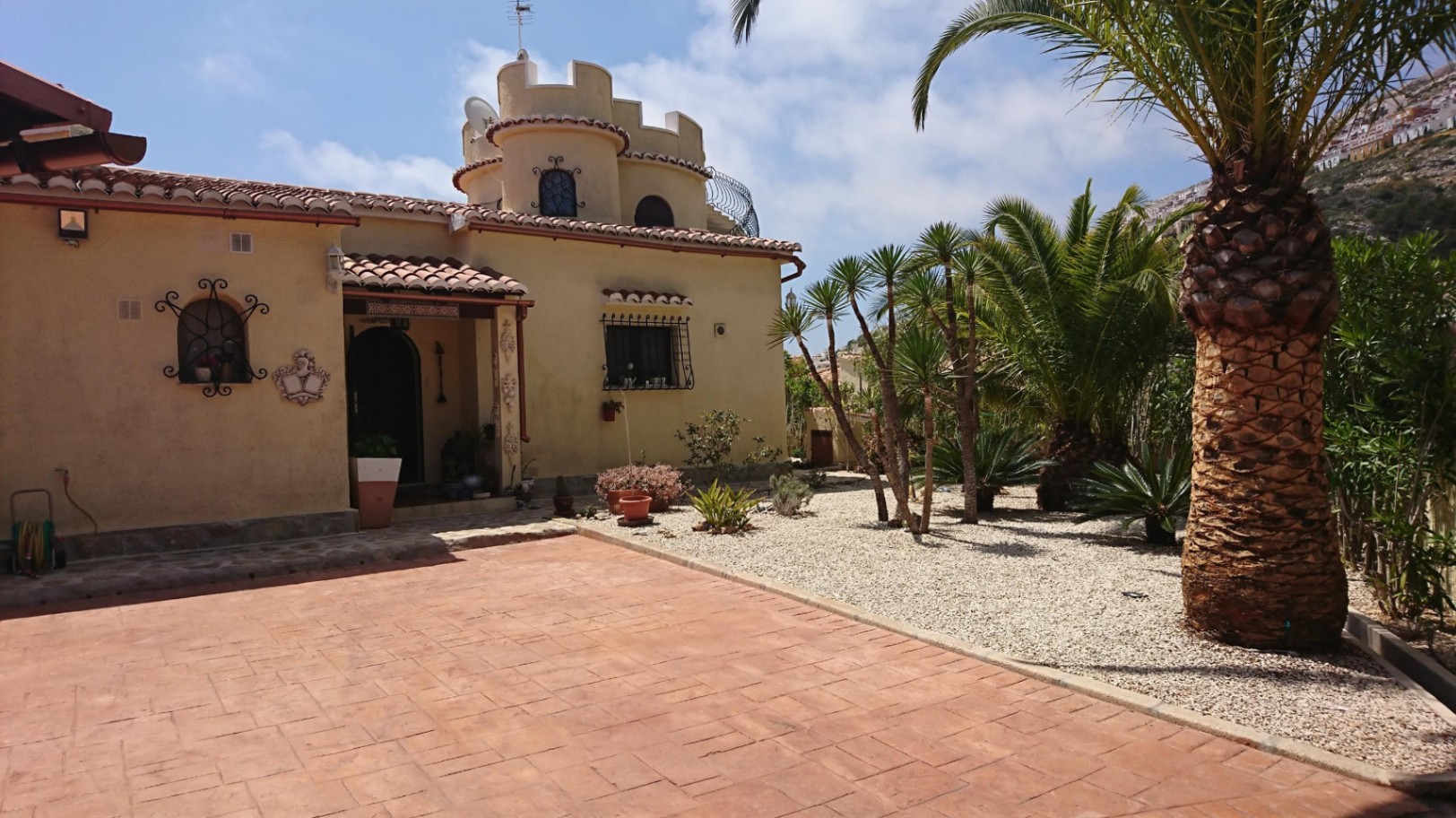 Außenansicht der Villa mit markantem Rundturm, Terrakottadach und einem mediterranen Kiesgarten mit Palmen und Sukkulenten.