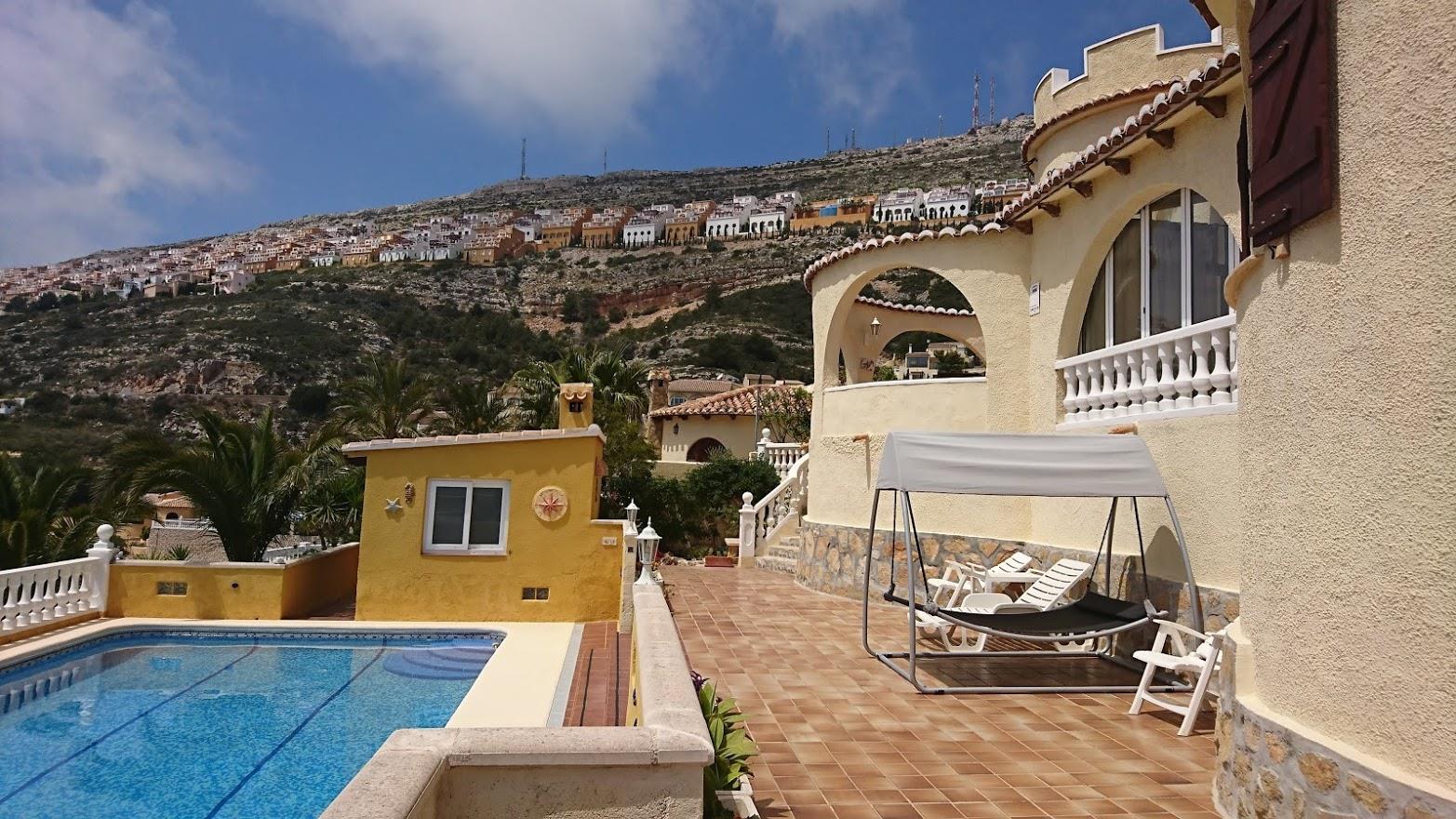 Außenansicht mit Swimmingpool, gefliester Terrasse und bogenförmiger Naya. Im Hintergrund erstreckt sich die Hügellandschaft der Urbanisation Cumbre del Sol.