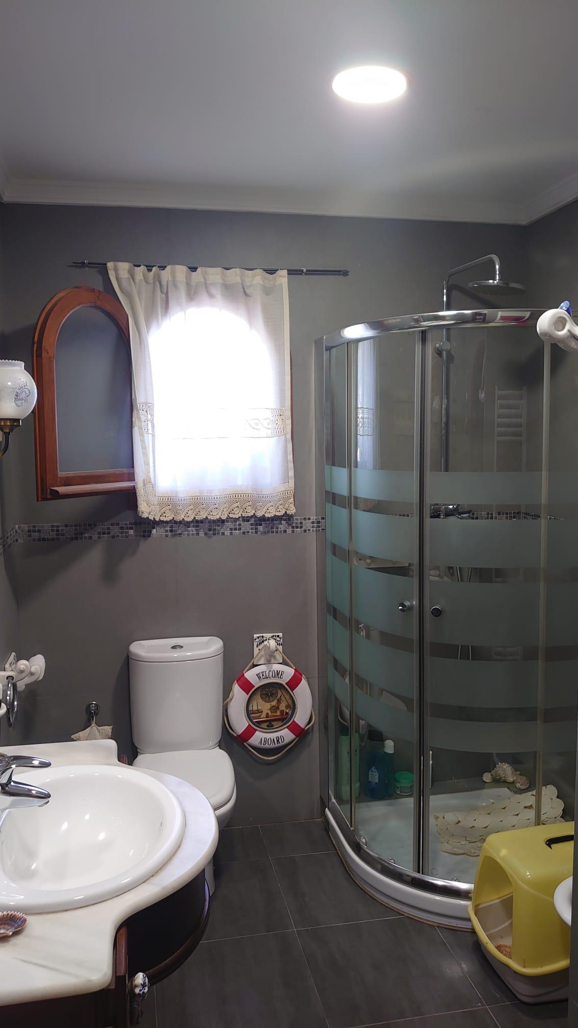 Badezimmer mit abgerundeter Glasduschkabine, grauen Fliesen, einem Rundbogenfenster aus Holz und maritimer Dekoration.