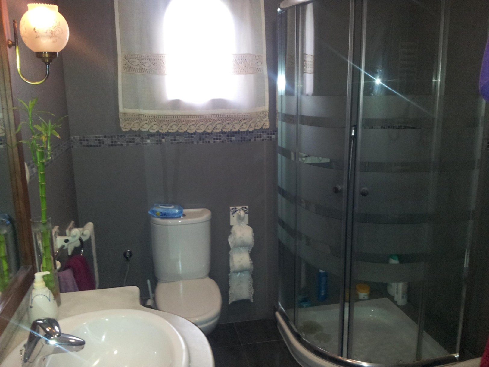 Badezimmer mit einer Eckduschkabine aus Glas, grauen Fliesen mit Mosaikbordüre und weißen Keramikarmaturen bei natürlichem Lichteinfall.