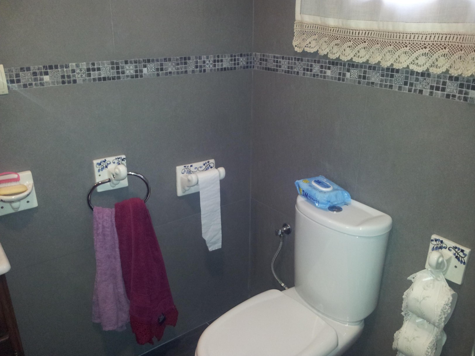 Badezimmer mit grauen Wandfliesen und Mosaikbordüre, ausgestattet mit weißem WC und traditionellen Keramik-Accessoires mit blauen Blumenmustern.