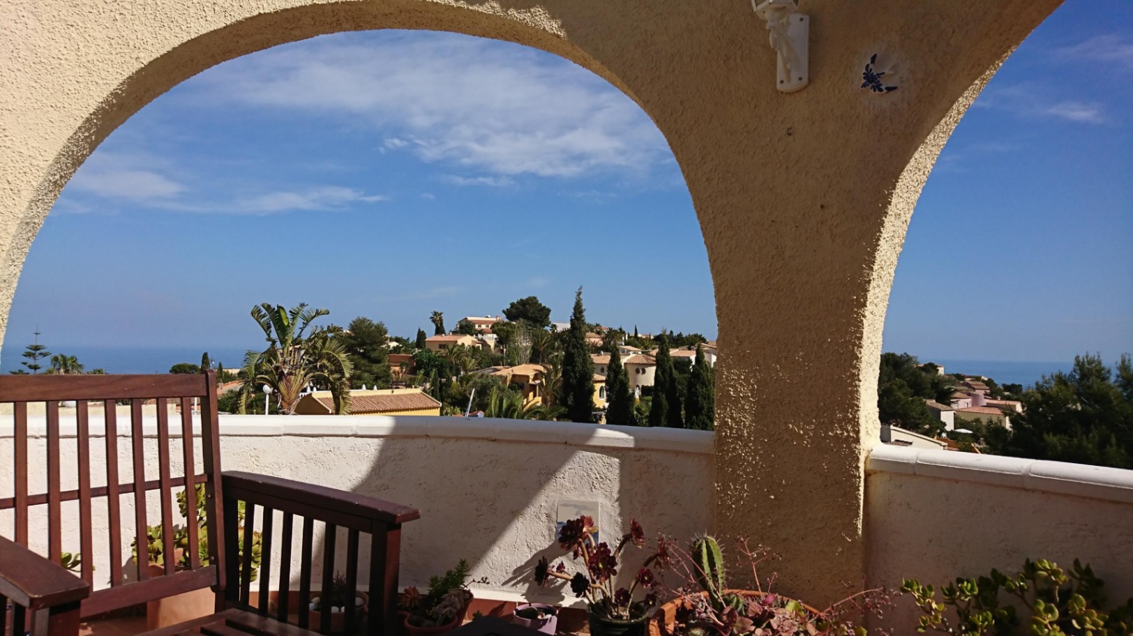 Blick von der bogenförmigen Naya auf das Mittelmeer und die umliegende Landschaft. Mit Holzmöbeln, Sukkulenten in Töpfen und traditioneller Architektur mit strukturiertem Putz.