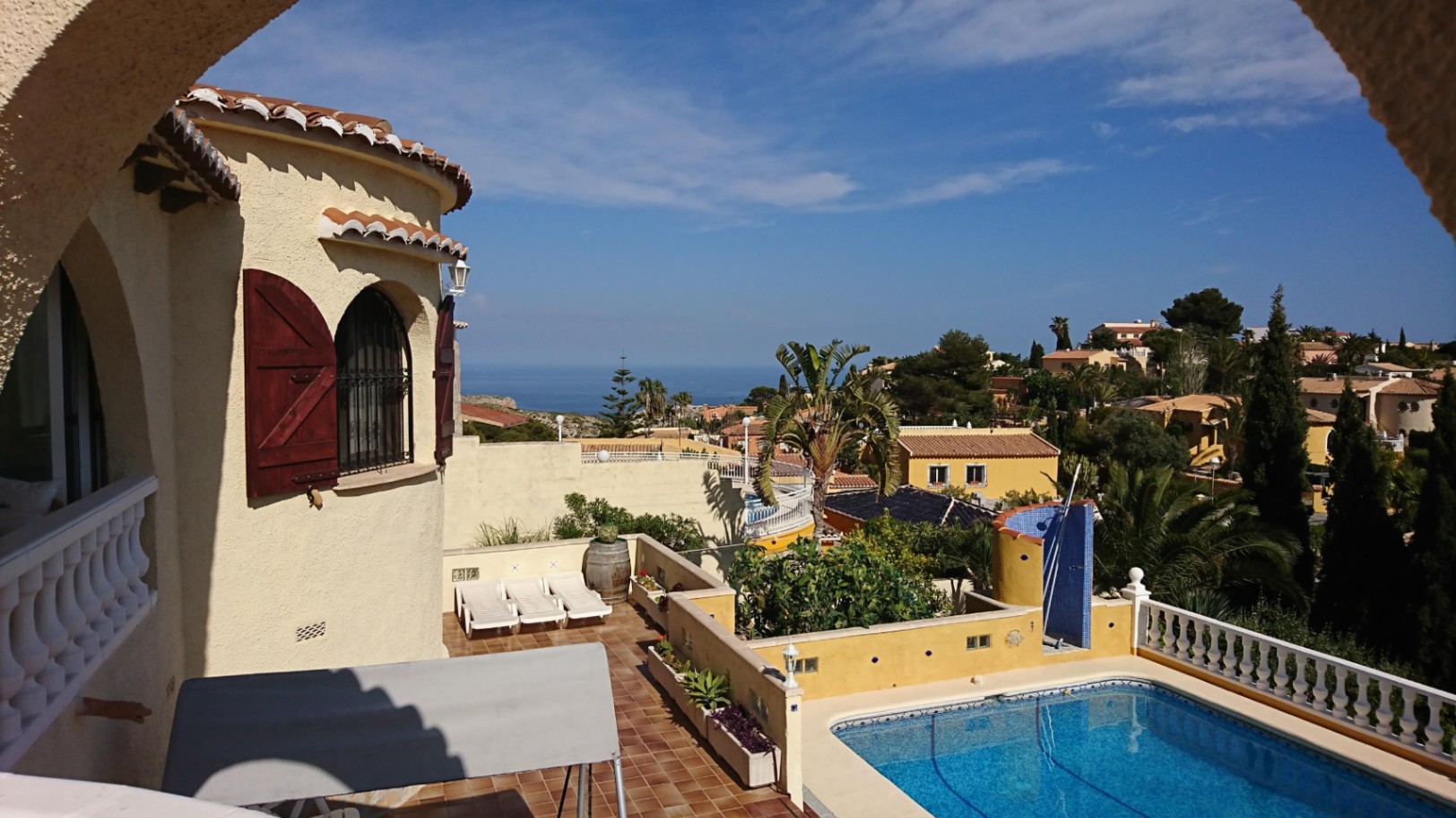 Erhöhte Ansicht des Außenbereichs mit Pool, Terracotta-Sonnenterrasse und Turmelement mit Holzläden, mit weitem Blick auf das Mittelmeer.