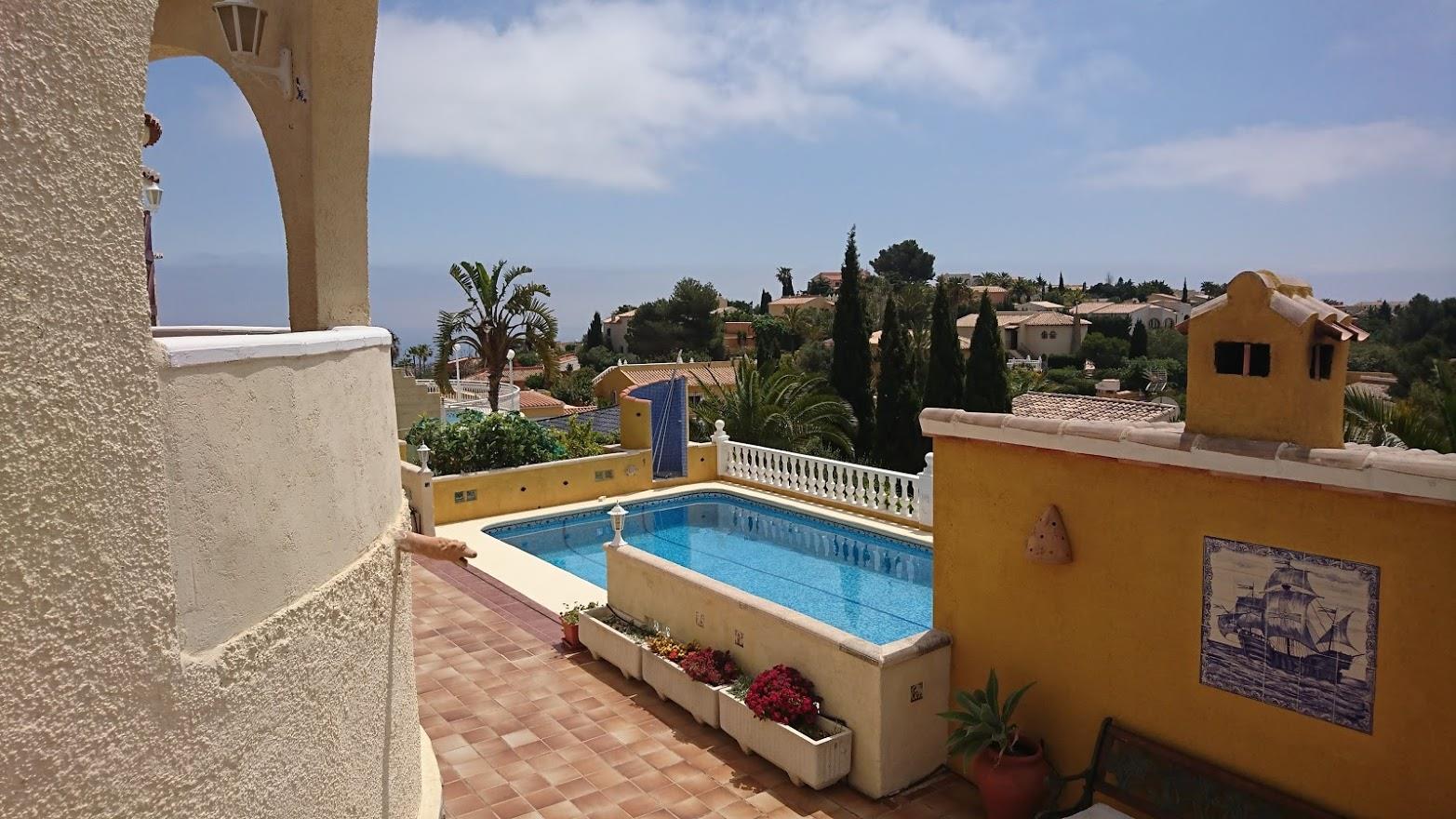 Erhöhter Blick auf den rechteckigen Pool mit Terrakotta-Terrasse, ockerfarbenen Mauern mit Zierfliesen und mediterraner Bepflanzung vor Meereskulisse.