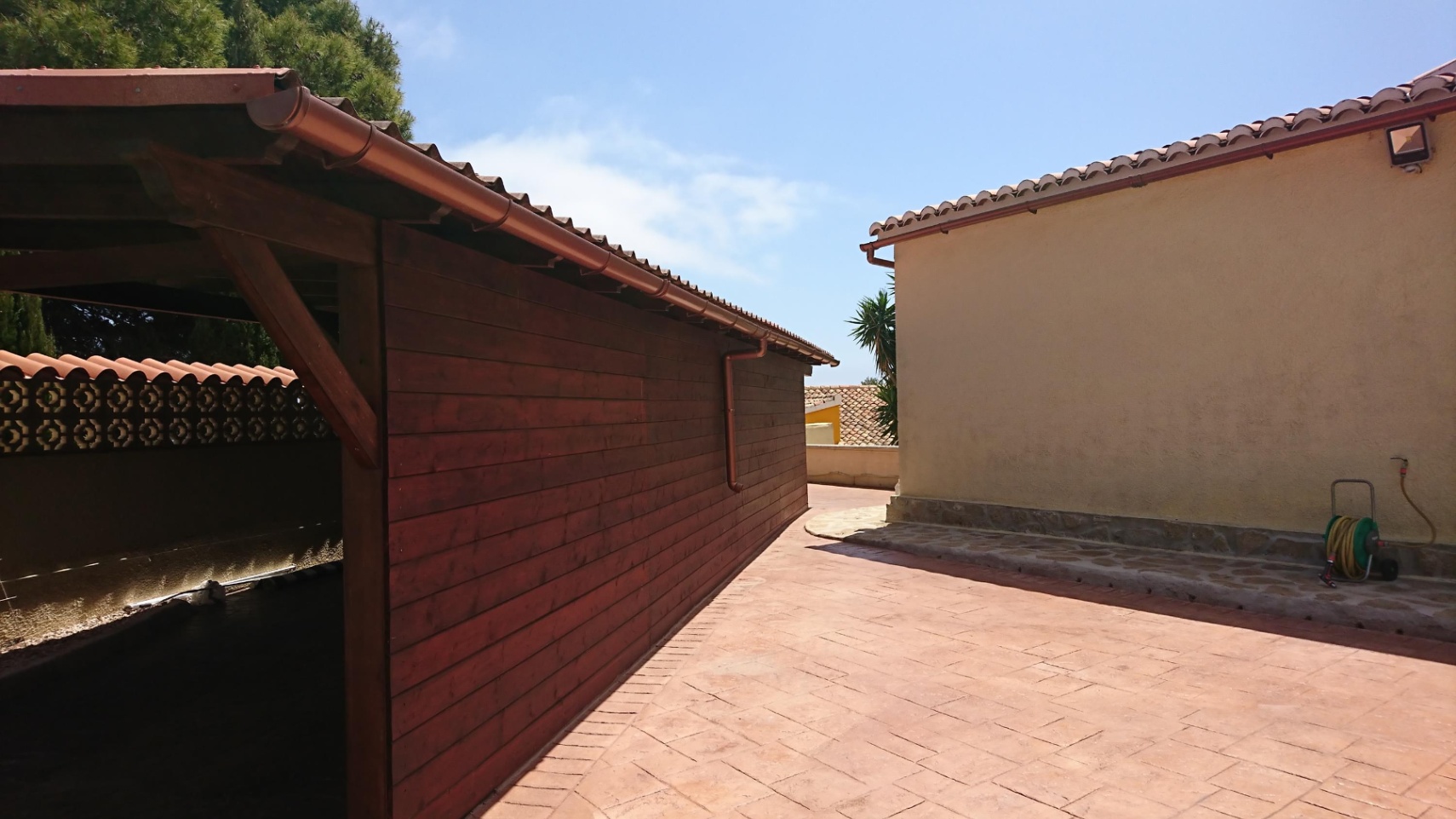 Gepflasterter Außenbereich mit einem Carport aus Holz und der Hausfassade, beide mit traditionellen mediterranen Ziegeldächern ausgestattet.