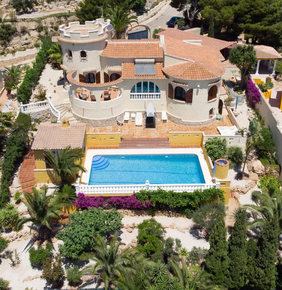 Luftaufnahme der mediterranen Villa mit Zinnenturm, einem 10x4m großen Pool und einem gepflegten Garten mit Palmen und Bougainvillea.