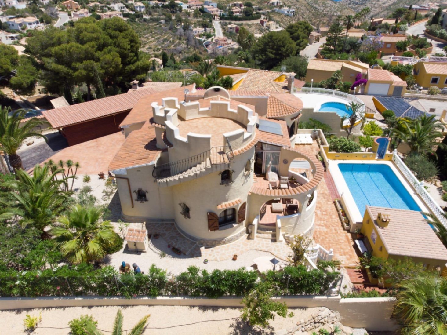Luftaufnahme der Villa mit markantem Rundturm, Dachterrasse, rechteckigem Pool und Außenküche, eingebettet in einen gepflegten mediterranen Garten.
