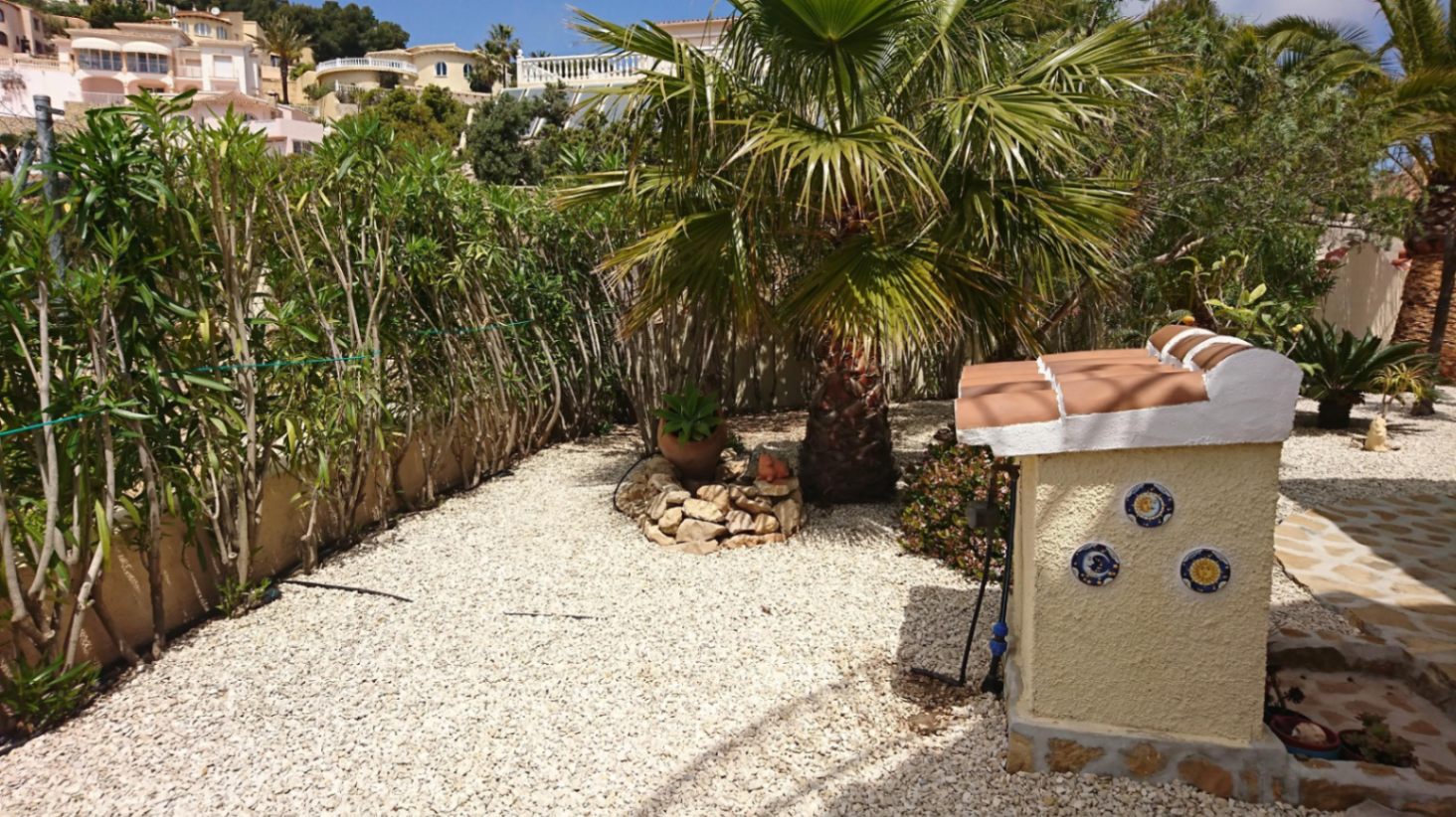 Pflegeleichter mediterraner Garten mit weißem Kies, Palmen und Hecken sowie einem dekorativen Häuschen mit traditionellen Fliesen.