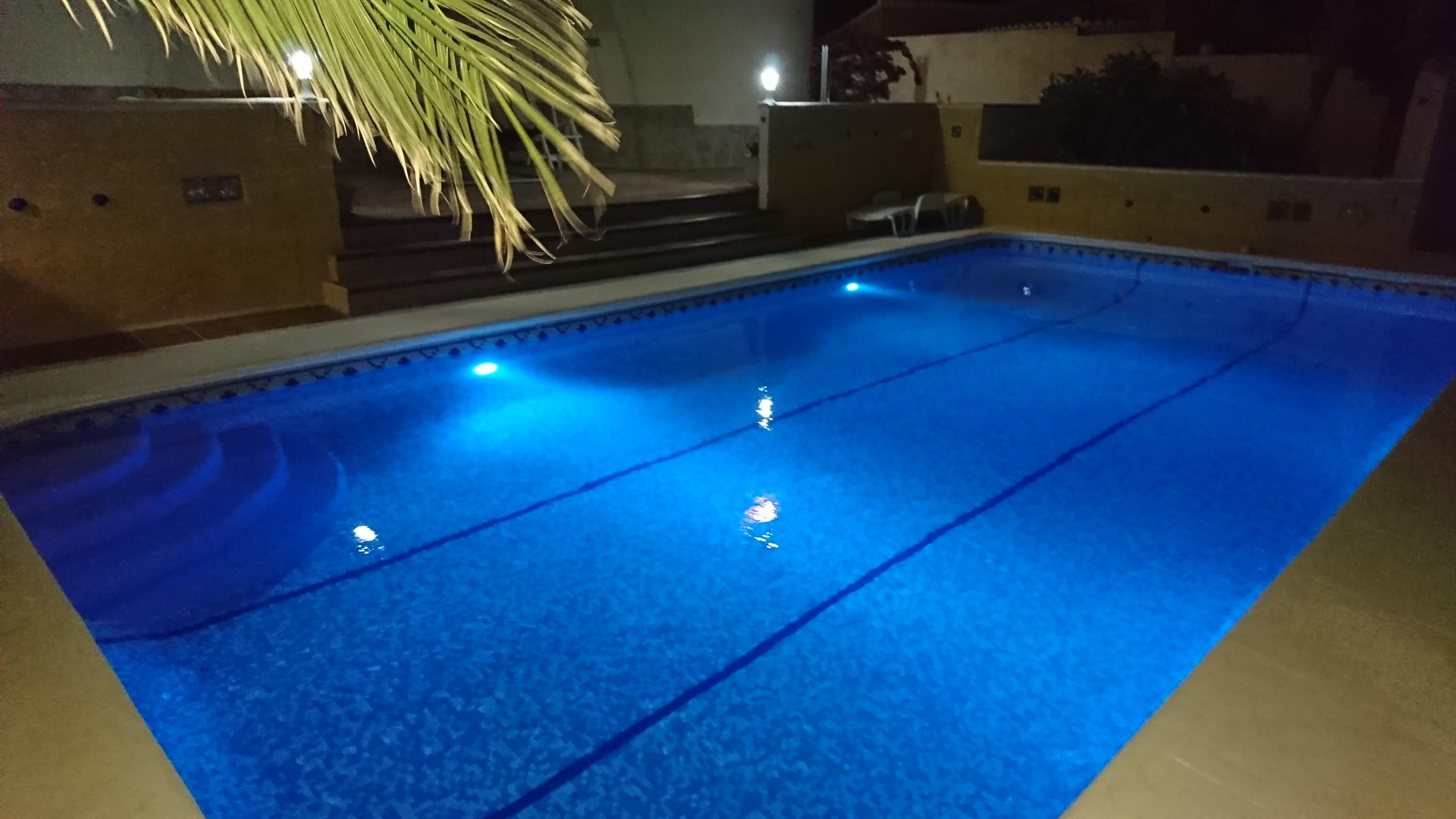 Rechteckiger Pool mit blauer Unterwasserbeleuchtung, römischer Treppe und gepflasterter Terrasse bei nächtlicher Außenbeleuchtung.