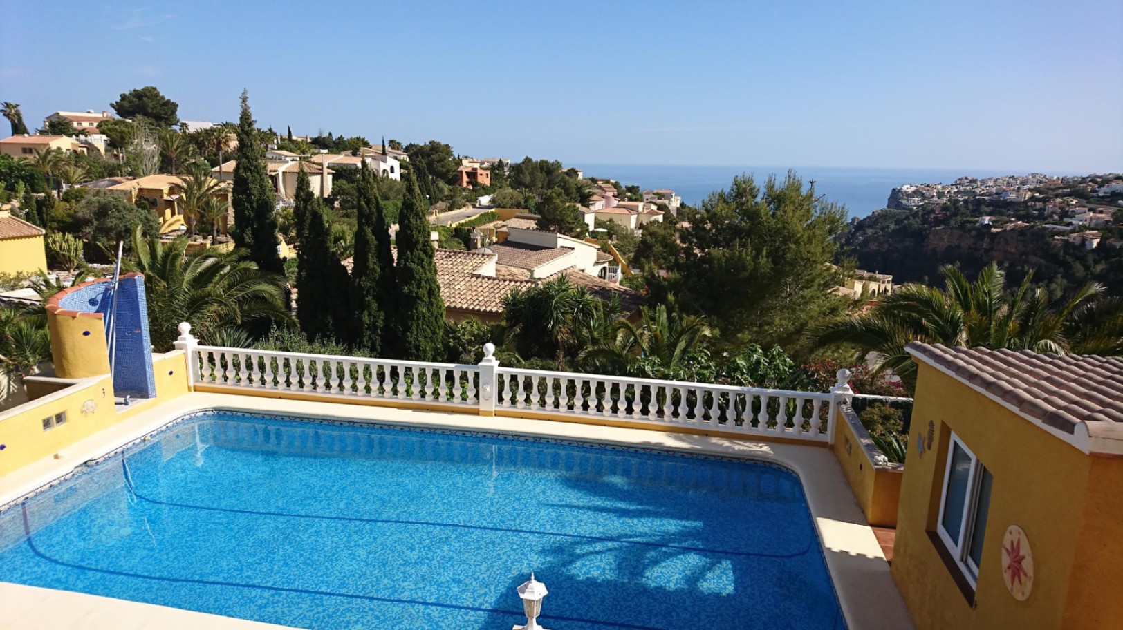 Rechteckiger Swimmingpool mit blauen Mosaikfliesen, weißer Balustrade und Panoramablick auf das Mittelmeer und die umliegende Küstenlandschaft.