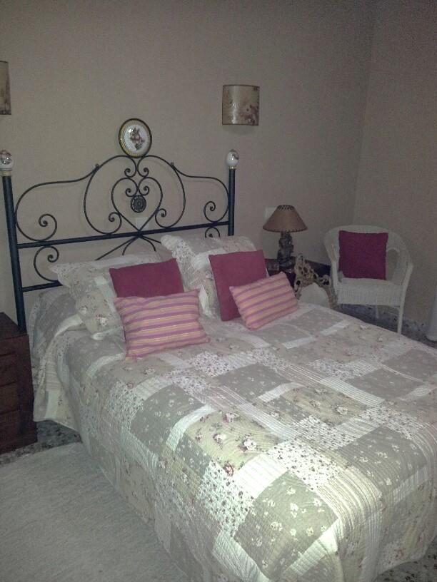 Schlafzimmer mit dekorativem schmiedeeisernem Kopfteil, Patchwork-Quilt und Korbsessel. Wandleuchten und eine klassische Nachttischlampe ergänzen die Einrichtung.