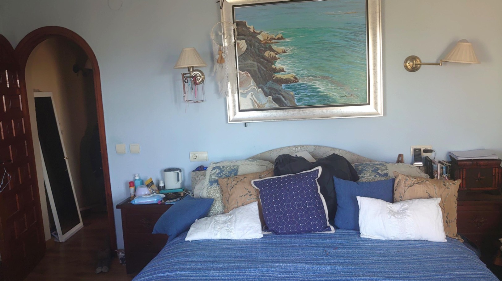 Schlafzimmer mit hellblauen Wänden, einem großen Küstengemälde über dem Bett, Wandlampen und einem markanten Rundbogen-Türrahmen aus Holz.