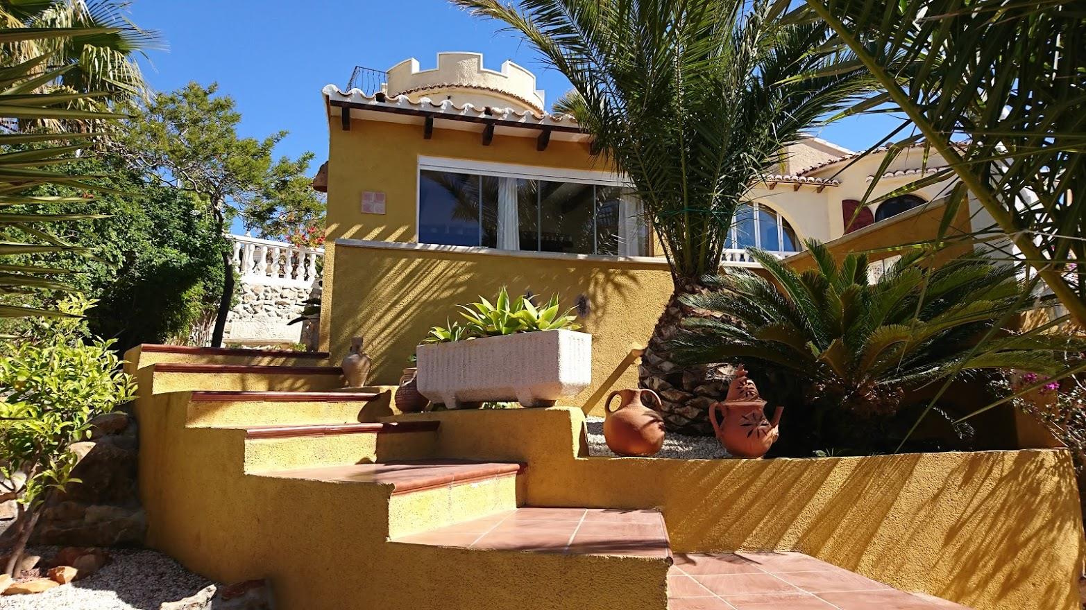 Terracotta-Außentreppen führen zur mediterranen Villa mit ihrem markanten Zinnenturm, ockerfarbenen Wänden und einem Garten mit Palmen und Amphoren.