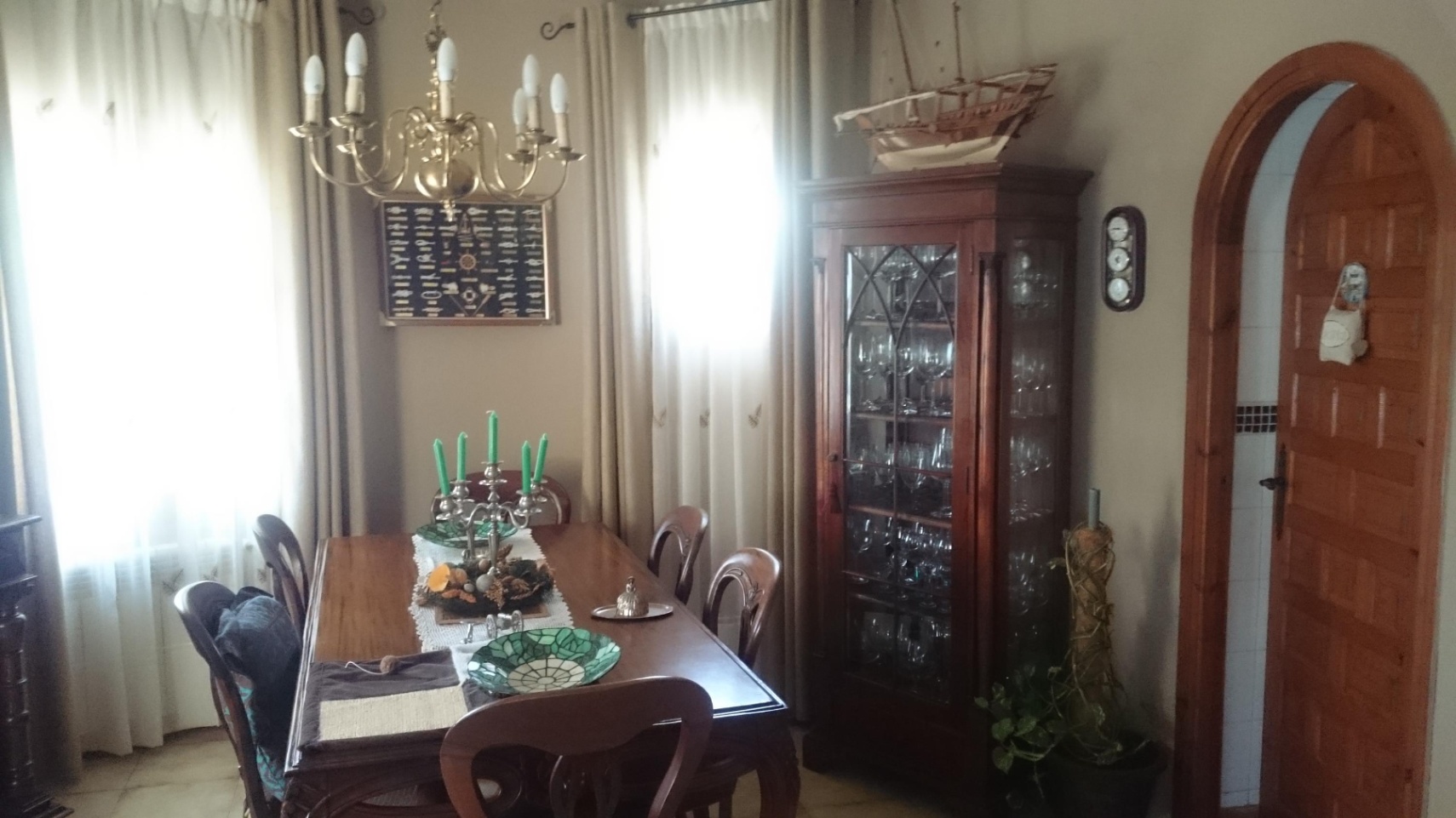 Traditionelles Esszimmer mit Holzmöbeln, einer Glasvitrine und einem Messingleuchter. Ein hölzerner Rundbogen und natürlicher Lichteinfall betonen den Raum.