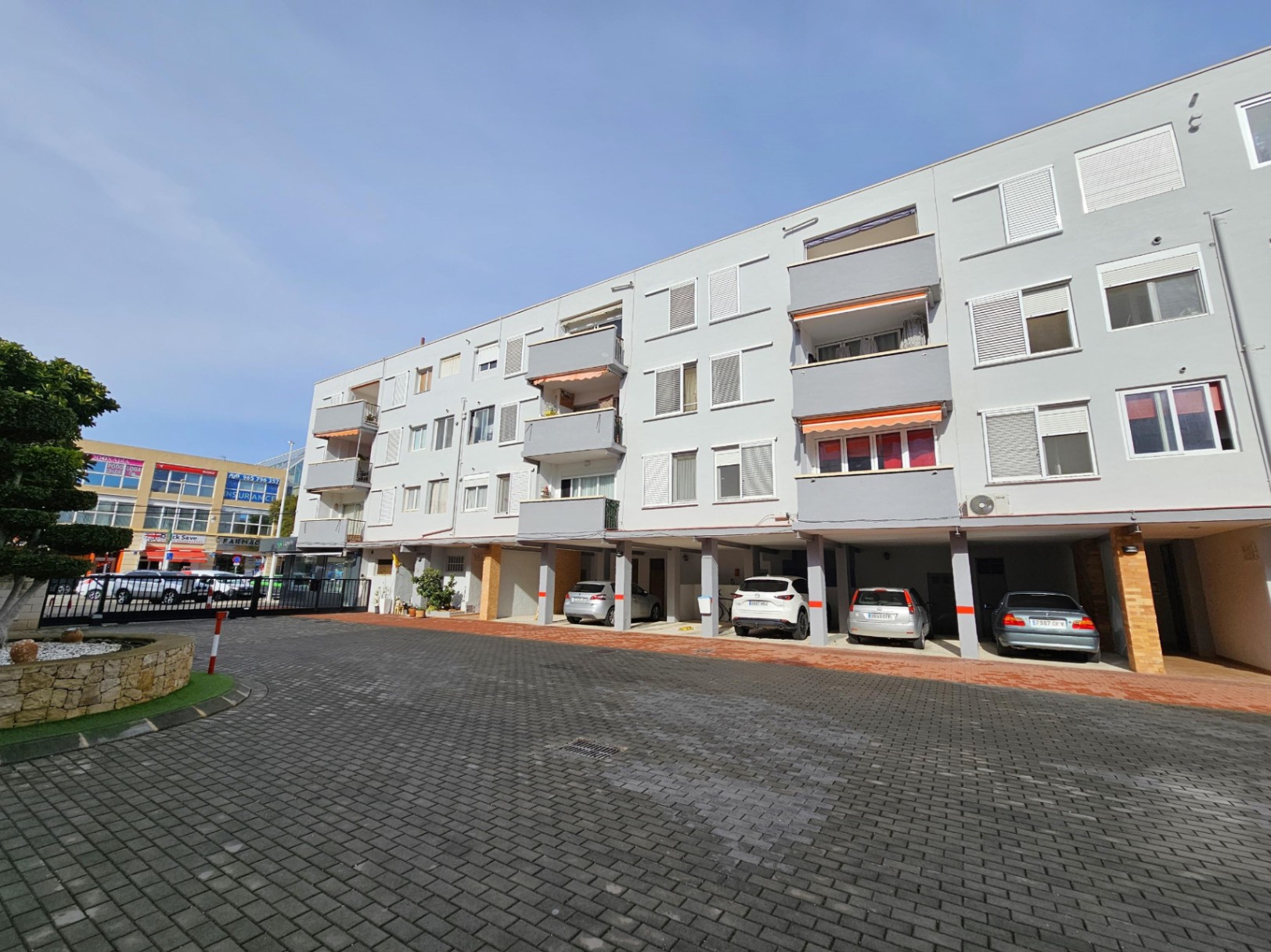 Außenfassade des Apartmenthauses mit privaten, überdachten Parkplätzen im Erdgeschoss und Balkonen mit Markisen über einem gepflasterten Hof.