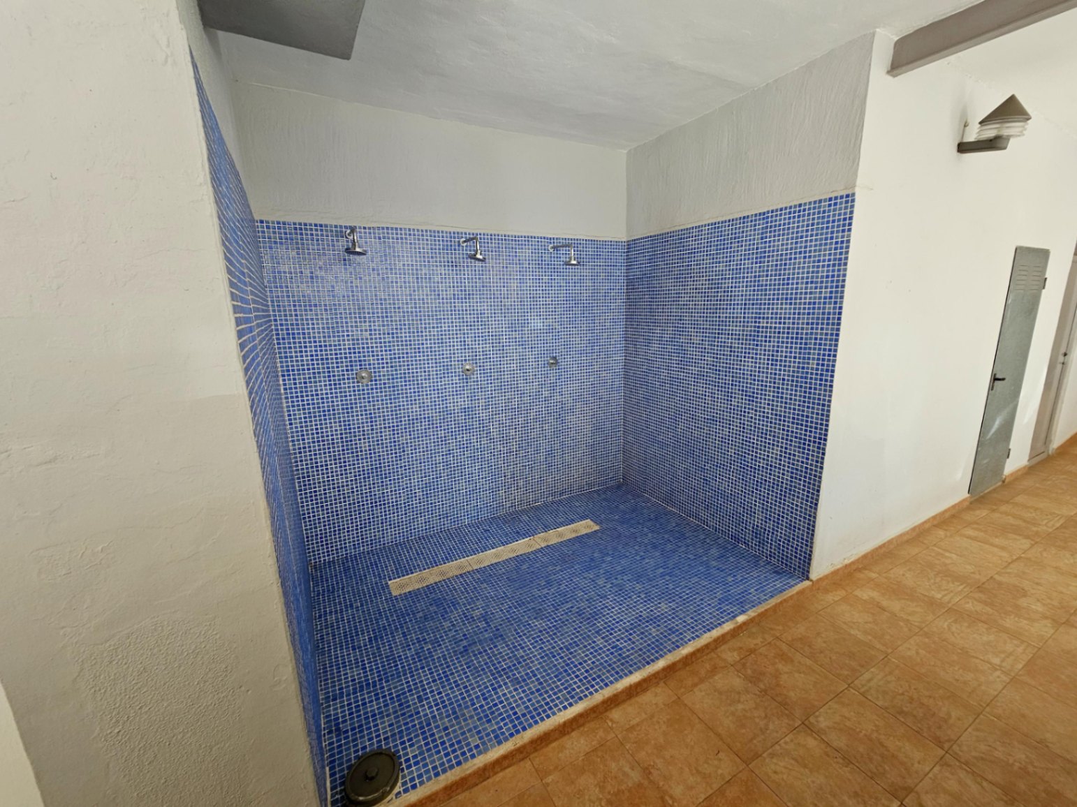 Gemeinschaftlicher Duschbereich mit blauen Mosaikfliesen, drei Duschköpfen und zentralem Abfluss in den Gemeinschaftsbereichen der Anlage.
